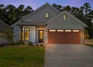 Modern single-story house with light gray siding, large front windows, a wooden front door, and a two-car garage with wooden doors. The porch light is on, and the house sits on a manicured lawn at dusk.