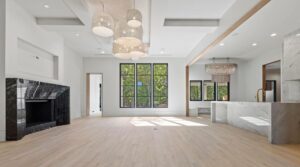 Spacious modern living area with light wood floors, large black-trimmed windows, a marble fireplace, pendant lights, and an open kitchen with a marble island and gold faucet. Natural light fills the room.