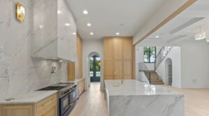 Modern kitchen with marble countertops and backsplash, light wood cabinets, a large island with gold faucet, stainless steel appliances, and an open layout leading to a staircase and arched doorway.