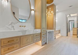 Modern bathroom with light wood cabinets, marble countertops, a large round mirror, gold fixtures, elegant lighting, and a vanity area. Light wood flooring and a hallway are visible in the background.