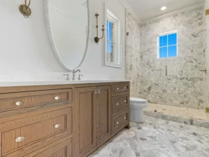 A bathroom with a wooden vanity, oval mirror, and marble countertop. The walk-in shower features marble walls and floors, a built-in bench, and a small window letting in natural light.