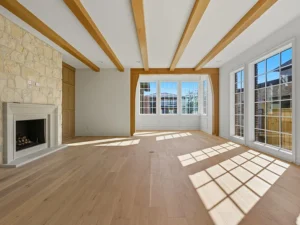 Bright, empty living room with wooden ceiling beams, light wood flooring, a stone fireplace on the left, and large windows on the right and back walls letting in natural sunlight.