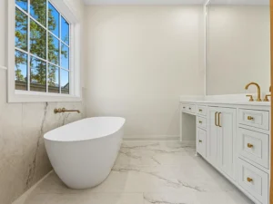 Modern bathroom with a freestanding white tub, gold fixtures, a large window with a view of trees, marble floor, and a white vanity with a large mirror and gold hardware.