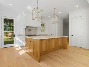 Modern kitchen with light wood floors, a large wooden island, white walls, marble countertops, gold pendant lights, a range hood, and a glass door leading outside.