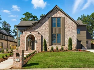 A two-story brick house with large windows, arched entryway, brown shutters, and manicured lawn. Tall trees and blue sky are visible in the background. A mailbox and pathway lead to the front door.