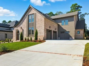 Modern two-story brick and wood house with steep rooflines, large windows, and a three-car garage. Neatly landscaped front yard with green grass, shrubs, and trees. Blue sky with scattered clouds overhead.