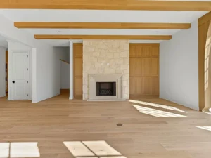A bright, empty living room with light wood floors, exposed wood ceiling beams, a central stone fireplace, and built-in wooden cabinets on either side. Sunlight streams through a window, casting shadows on the floor.