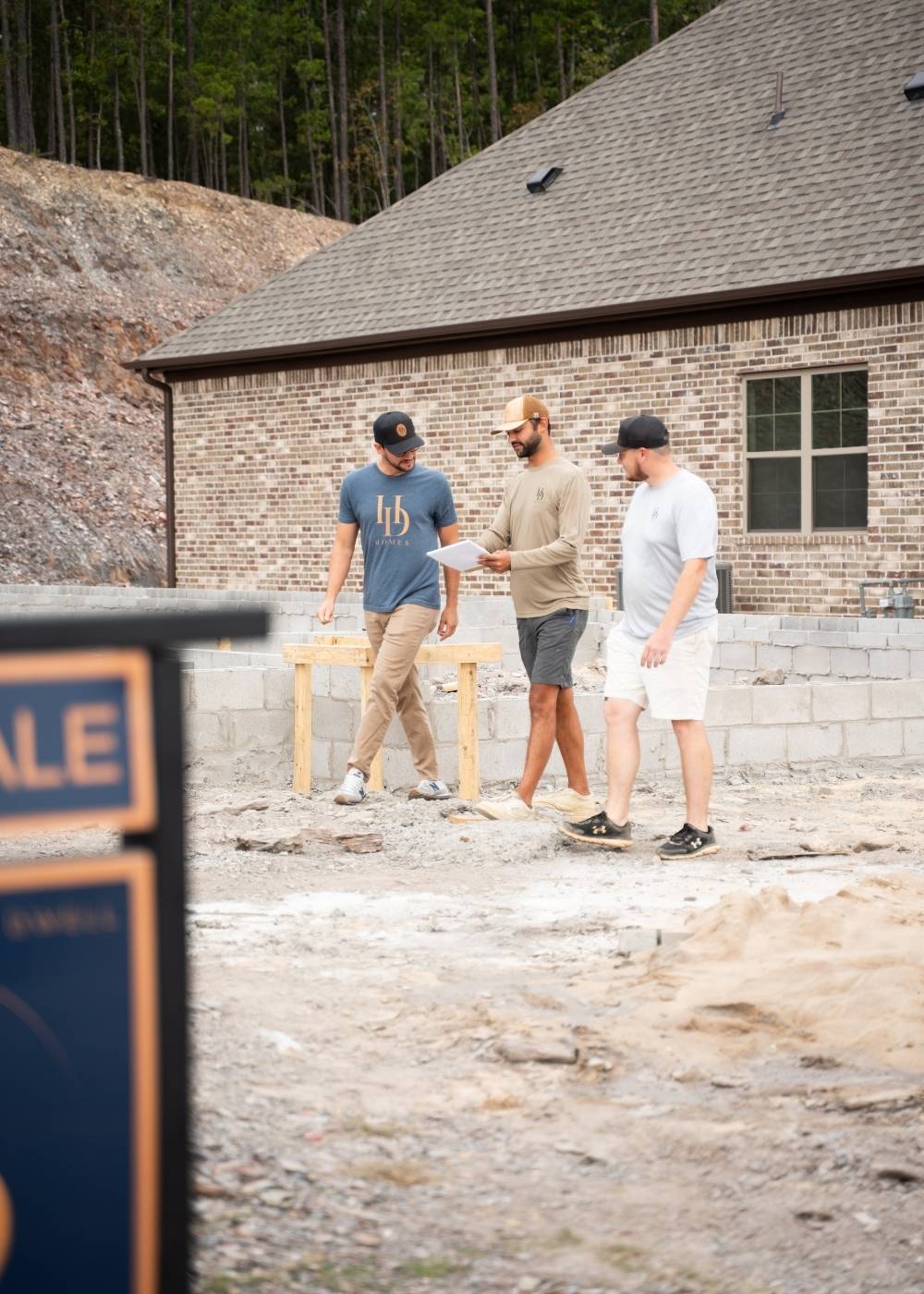 Three men wearing casual clothes and caps walk and talk at a construction site in front of a brick house. A For Sale sign is visible in the foreground. Trees and dirt are in the background.