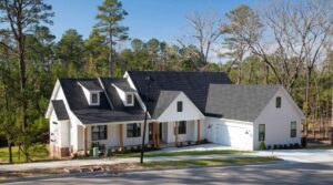 A modern white house with black roof shingles, large windows, and a front porch sits near a forested area. There is a driveway leading to a double garage and a well-kept lawn bordered by trees.