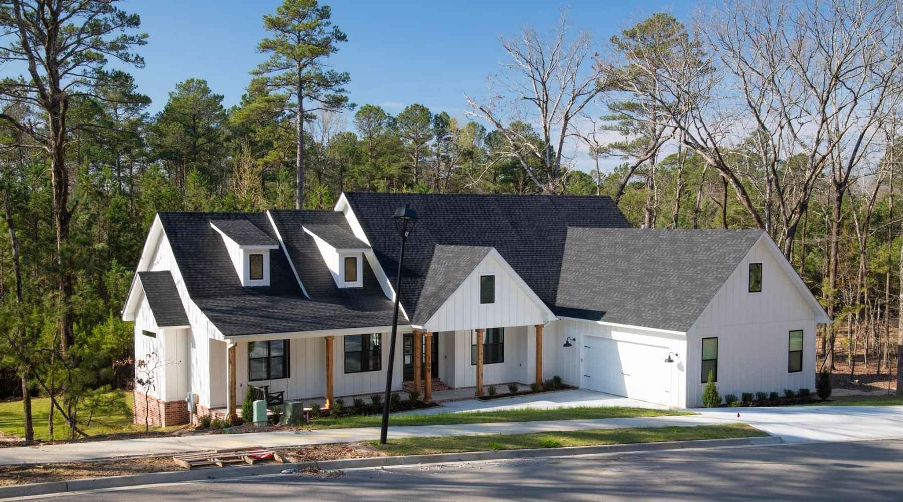 A modern white house with black roof shingles, large windows, and a front porch sits near a forested area. There is a driveway leading to a double garage and a well-kept lawn bordered by trees.