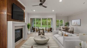 A modern living room with a white sofa, round coffee table, wall-mounted TV above a fireplace, wooden accent wall, large windows, sliding doors to a patio, and a dining area in the background.