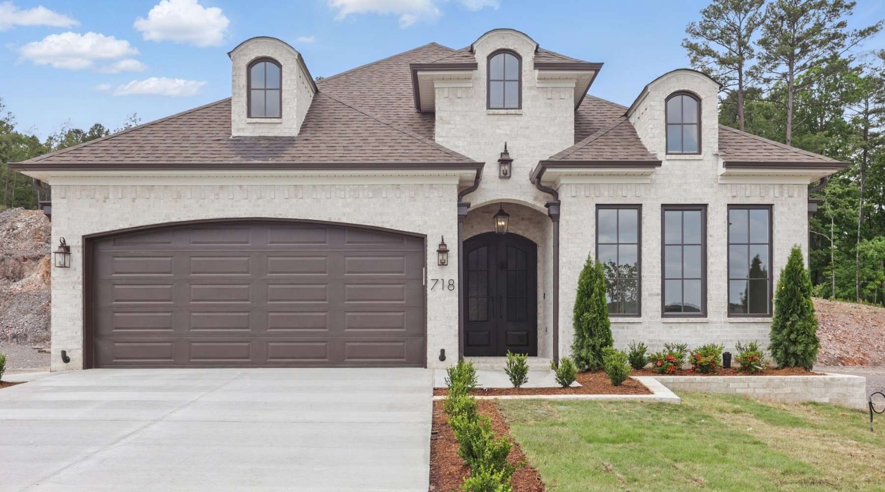A modern two-story house with light brick exterior, dark brown garage door, and black-trimmed windows. Manicured landscaping lines the front walkway, and the house sits on a concrete driveway.