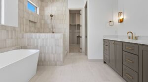 Modern bathroom with a freestanding white bathtub, walk-in shower with beige tiles, gray vanity with gold fixtures, wall-mounted light, and a closet visible through an open door in the background.