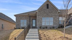 A single-story brick house with large windows, a steeply pitched roof, and a central front door. Concrete steps with a black railing lead up through a dry, grassy yard to the entrance.