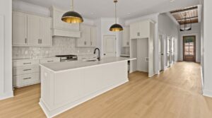 Modern kitchen with white cabinets, a large island with a sink, light wood floors, two black-and-gold pendant lights, stainless steel appliances, and a hallway leading to a glass door in the background.