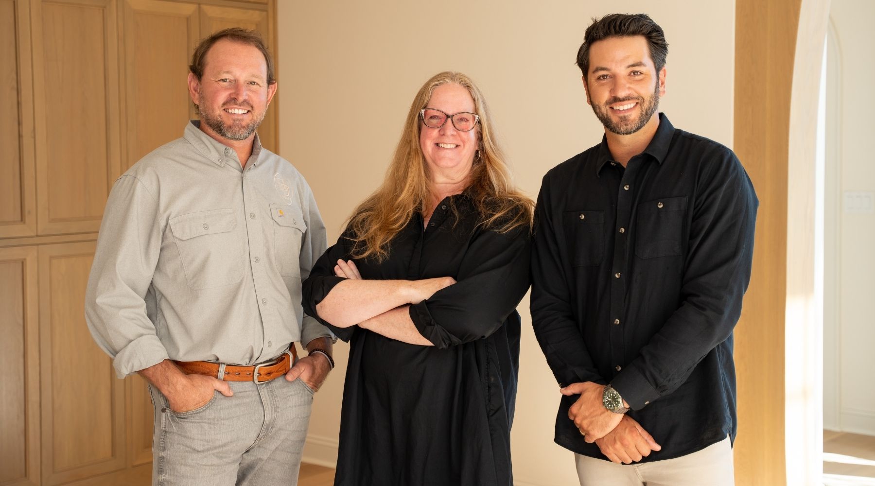 Three people stand indoors, smiling at the camera. The two men wear button-up shirts and the woman in the center wears glasses and a black dress with her arms folded. Light wood cabinets and a sunlit archway are visible in the background.