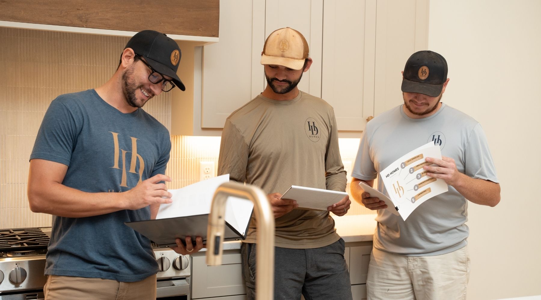 Three men in casual branded shirts and hats stand in a modern kitchen, reviewing documents and a tablet together, appearing focused and engaged in discussion.