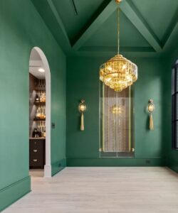 A modern room with green walls, a gold chandelier, two wall sconces, light wood floors, and an arched doorway leading to a small bar area with shelves of bottles. Large windows let in natural light.