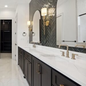 Modern bathroom with a double vanity, white countertops, black cabinets with gold handles, large arched mirror, gold faucets, and wall sconces. The floor and walls are white marble, and there is a walk-in closet visible.