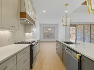 Modern kitchen with light gray cabinets, a gas stove, white countertops, and gold pendant lights over an island with a sink and dishwasher. Large windows and glass doors let in natural light.