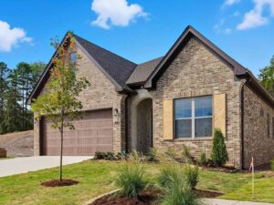 A modern one-story brick house with a two-car garage, wooden shutters, arched entryway, and well-maintained lawn with young trees and shrubs, set against a blue sky with a few clouds.