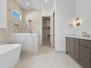 Modern bathroom with a freestanding white tub, a walk-in shower with beige tiles, gray double-sink vanity with gold fixtures, wall sconce, and a walk-in closet seen through an open door. Neutral color scheme and bright lighting.