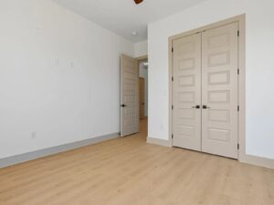 A bright, empty room with light wood flooring, white walls, double beige closet doors, and an open beige door leading to a hallway.