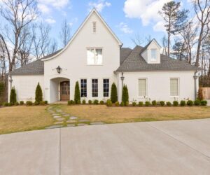 A large, modern white brick house with a steep roof, dormer windows, and a front yard featuring a stone path, trimmed bushes, and a wide driveway; trees and blue sky in the background.