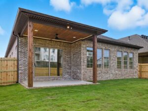 A covered backyard patio with a wooden ceiling, ceiling fan, and brick exterior wall, next to a green lawn and fenced yard, attached to a single-story house with large windows.