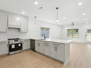 Modern kitchen with light gray cabinets, white countertops, stainless steel stove, large island with sink, wood flooring, pendant lights, and open layout leading to living area with large windows.