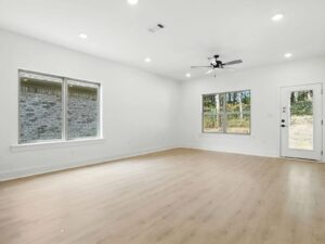 A spacious, empty room with light wood flooring, white walls, two large windows, a ceiling fan with lights, and a door with a glass panel letting in natural light.