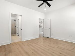 A bright, empty room with light wood flooring, white walls, a ceiling fan, and three open doors leading to a bathroom, a hallway, and a kitchen area.