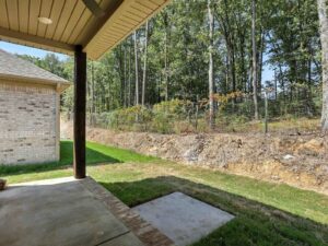 A covered patio with a concrete floor overlooks a grassy backyard bordered by a wire fence and trees, with a house on the left and a clear, sunny sky above.
