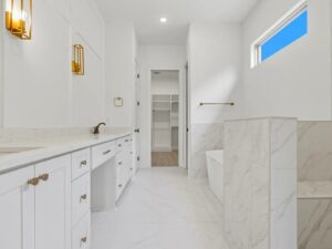 A modern white bathroom with marble floors and walls, double sinks with gold fixtures, a built-in bathtub, and a walk-in closet at the back. Natural light enters through a high window.
