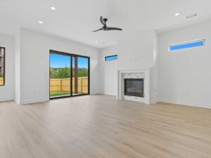 Bright, modern living room with light wood floors, white walls, large sliding glass doors to a fenced yard, a ceiling fan, and a fireplace with a white mantel and tile surround.