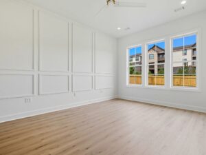 Bright, empty room with white paneled walls, light wood flooring, a ceiling fan, and large windows showing a view of a wooden fence and neighboring buildings under a blue sky.