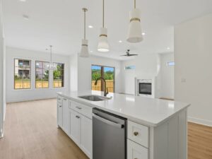 Modern, bright kitchen with white cabinets, a large island with sink and dishwasher, pendant lights, and open view to living area with fireplace. Windows and glass door provide natural light and views of a fenced backyard.