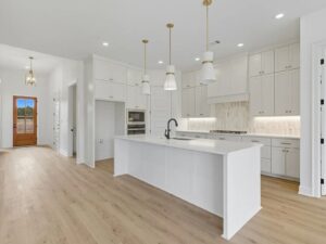 Modern kitchen with white cabinets, a large island with a sink, built-in oven, gas cooktop, pendant lights, light wood floors, and recessed lighting. A hallway and wooden door are visible in the background.