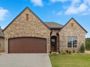Single-story brick house with a steep gable roof, brown double garage doors, an arched front entrance, and a neatly landscaped front yard under a bright blue sky with scattered clouds.