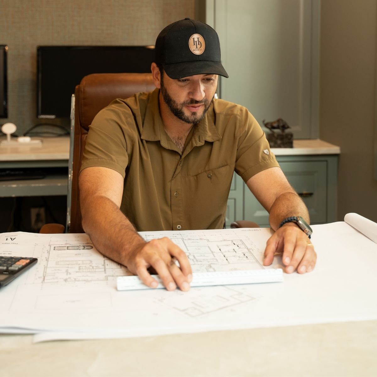 A man in a brown shirt and black cap studies architectural blueprints at a desk, using a ruler to measure. Office equipment and a computer are visible in the background.