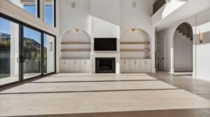 A bright, modern living room with large windows, built-in shelves and cabinets flanking a fireplace with a mounted TV above. The space has light wood floors, white walls, and an open staircase on the right.