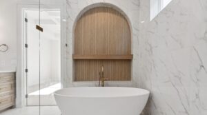 A modern bathroom with white marble walls, a freestanding oval bathtub, a gold faucet, and a niche with wood paneling and a shelf above the tub. Natural light enters through a window, creating a bright atmosphere.