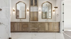 Double vanity bathroom with two mirrors, tall cabinets, light wood cabinetry, marble floor, wall sconces, and glass shower doors. Reflections show a bright, modern, and spacious design.