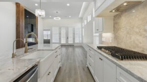 Modern kitchen with white cabinets, marble countertops, stainless steel fixtures, a gas stovetop, and a farmhouse sink. Light wood flooring and large windows bring in natural light from the adjacent open living area.