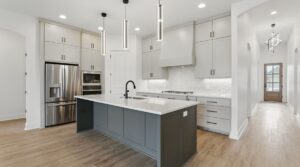 Modern kitchen with light gray cabinets, stainless steel appliances, a large dark gray island with a white countertop, pendant lights above, wood floors, and a hallway leading to a door with glass panes.