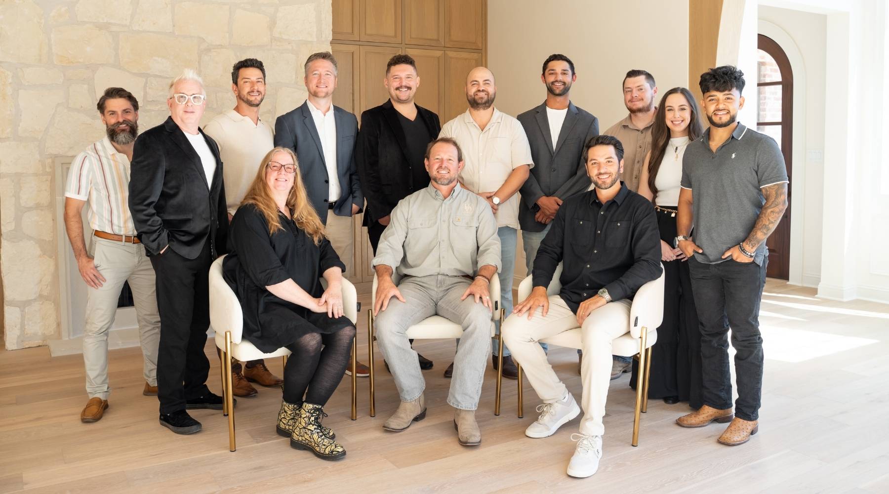 A group of thirteen people, dressed in business casual attire, pose together in a bright room with wooden floors and a stone wall. Some are seated while others stand behind them, all smiling at the camera.