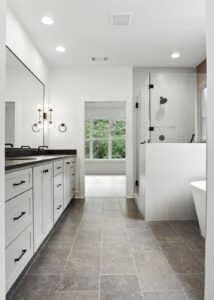 Modern bathroom with gray tiled floor, double sink vanity with white cabinets, large mirror, glass-enclosed shower, freestanding bathtub, and a large window letting in natural light.