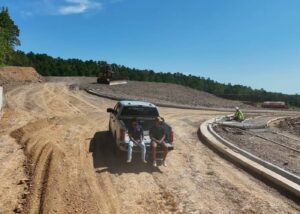 Two people sit on the tailgate of a white pickup truck parked on a dirt construction site, while a worker in a safety vest works nearby and a yellow excavator is visible in the background under a clear blue sky.