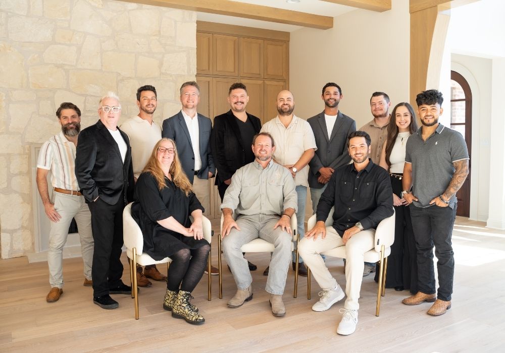 A group of fourteen adults, consisting of eleven men and three women, pose indoors in a well-lit room. Three people are seated in front while the rest stand behind them, all smiling at the camera.