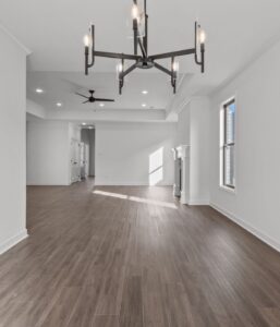 Modern, empty living space with wood flooring, white walls, large windows, a black chandelier, ceiling fan, and a fireplace. Natural light streams in, creating a bright, airy atmosphere.